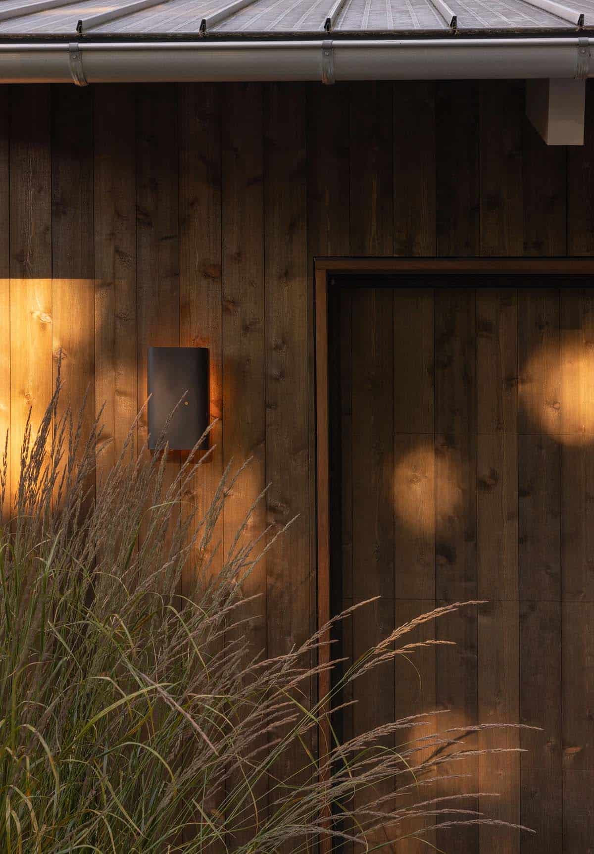 Exterior dusk detail of dark vertical wood siding with a minimalist wall sconce casting warm light
