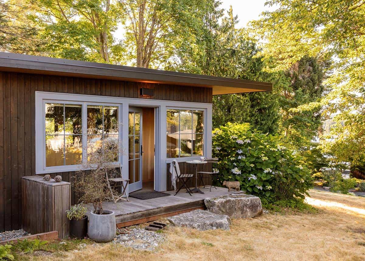 Small studio outbuilding with dark wood siding, French doors, small deck, and surrounding hydrangeas and mature trees