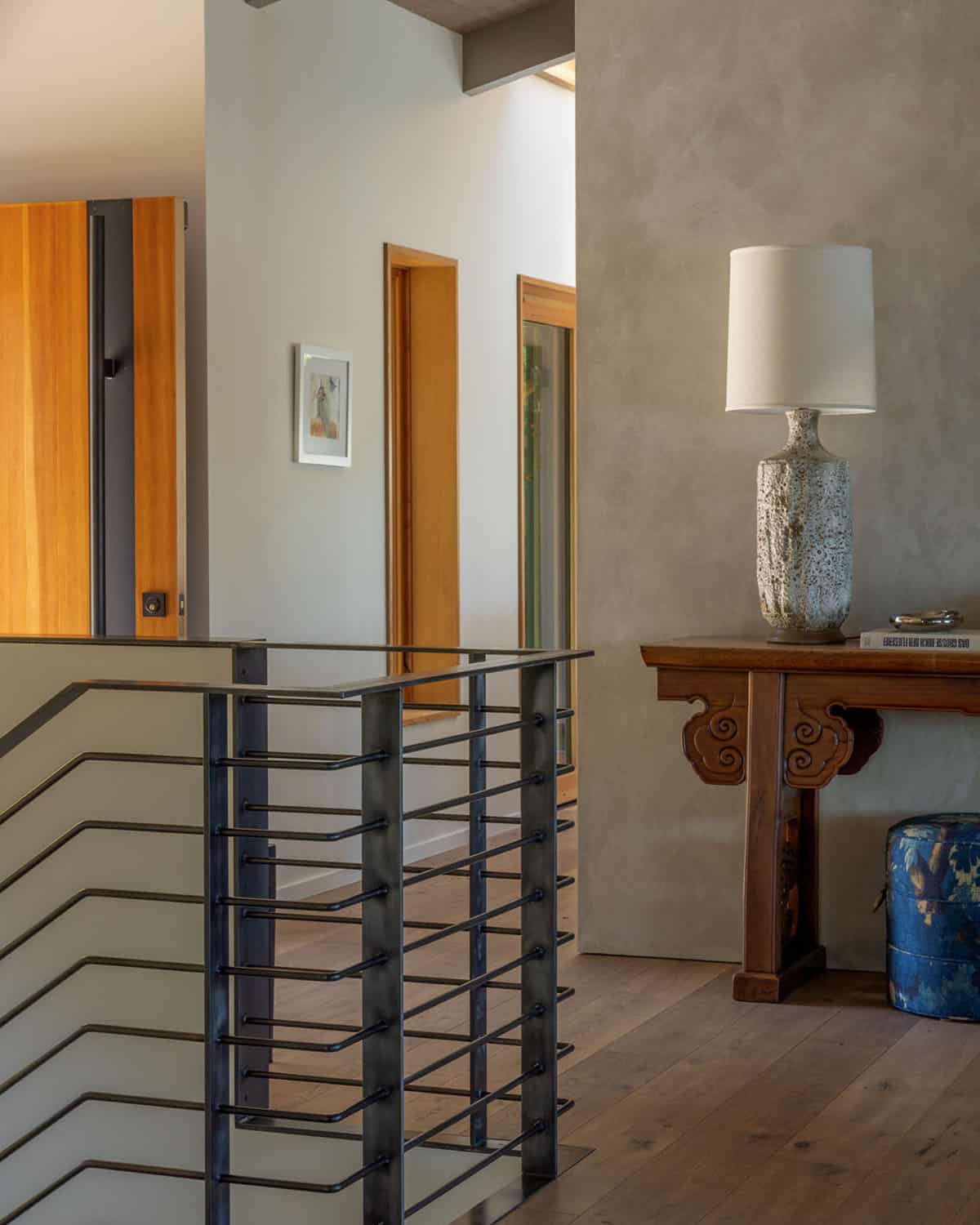 Upper landing with a custom steel railing, textured plaster walls, warm wood door frames, and an antique Chinese console table