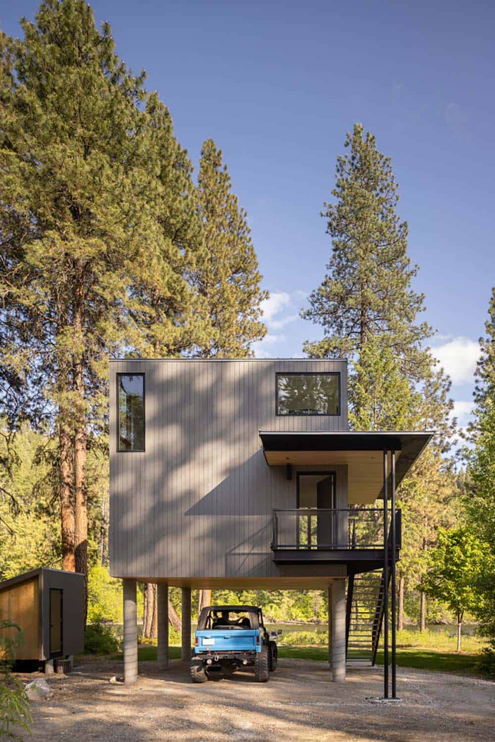 Modern cabin on concrete stilts with vertical wood siding and blue Jeep below