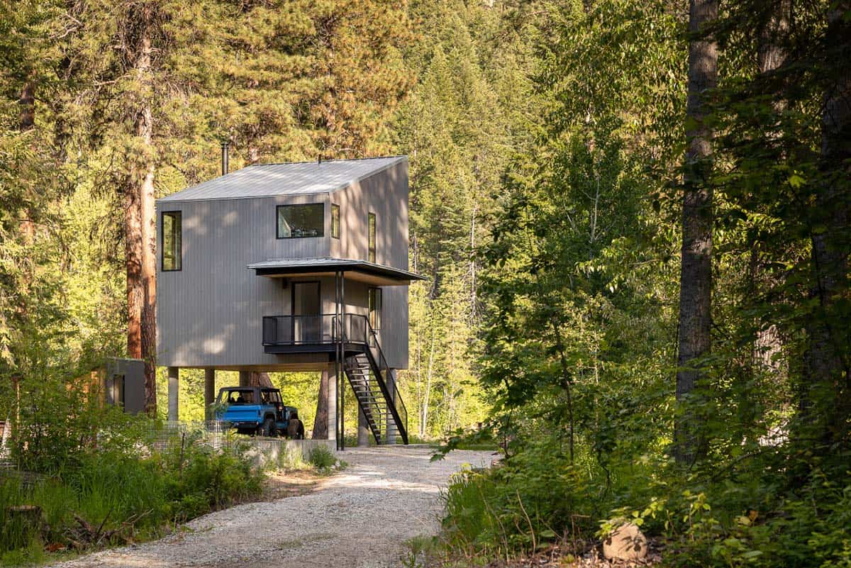 Modern elevated cabin on concrete stilts viewed from forest path with blue Jeep