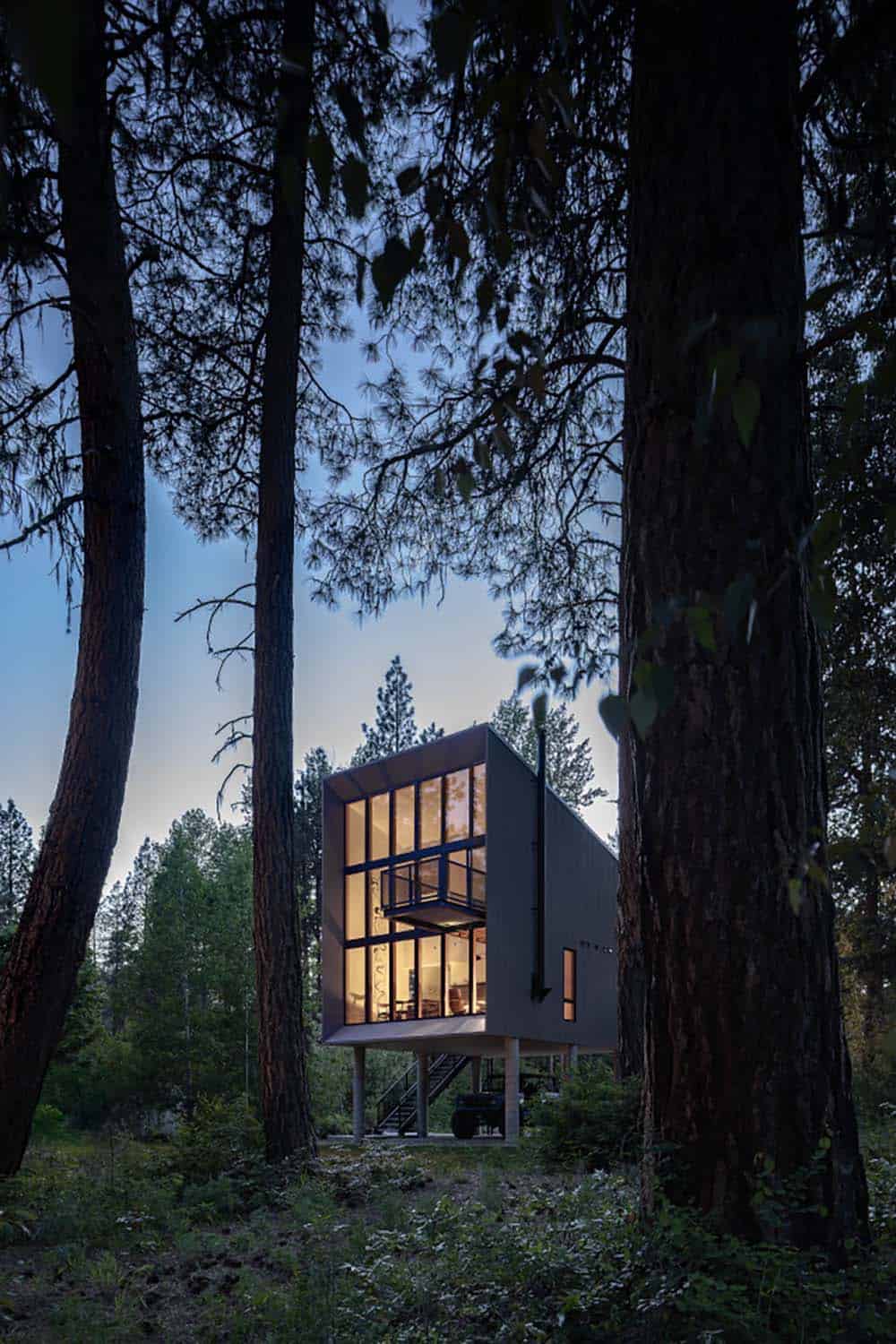 Modern cabin glowing at dusk seen through dark pine forest Washington