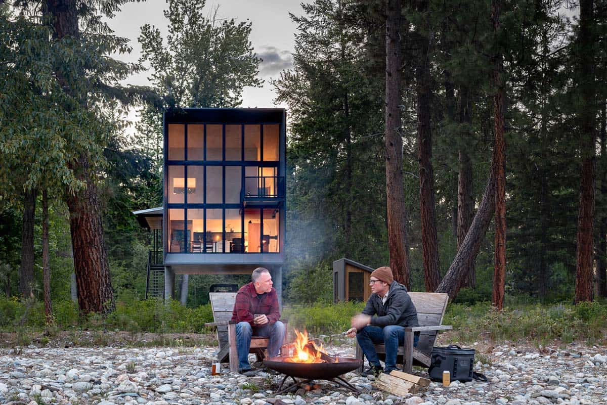 Two men sitting by campfire in front of glowing modern cabin on Wenatchee River