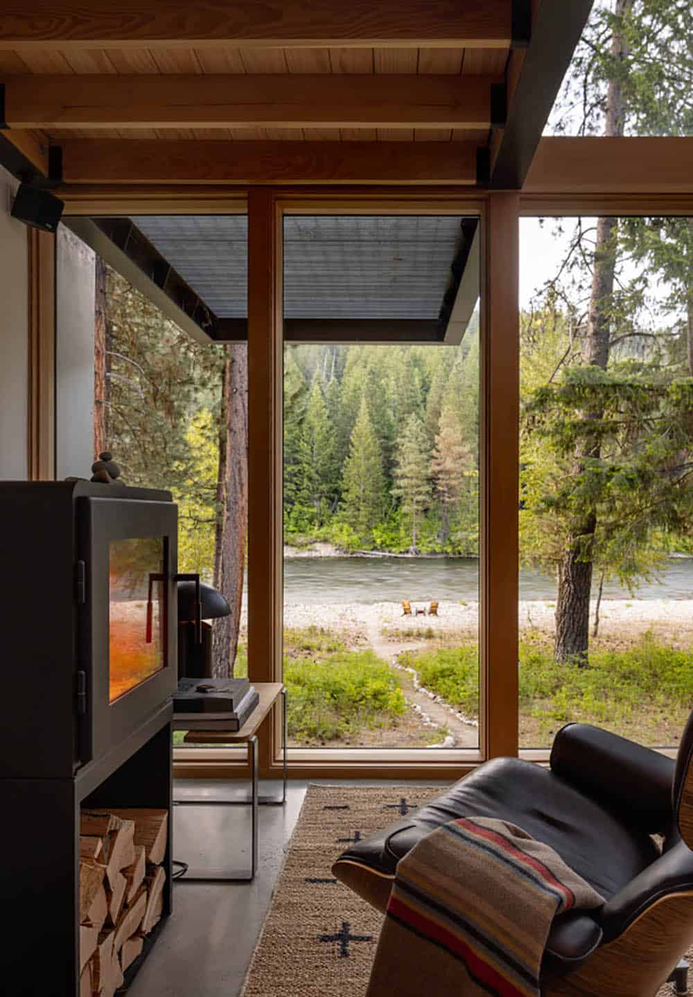 Eames lounge chair by wood stove with floor-to-ceiling river and forest views