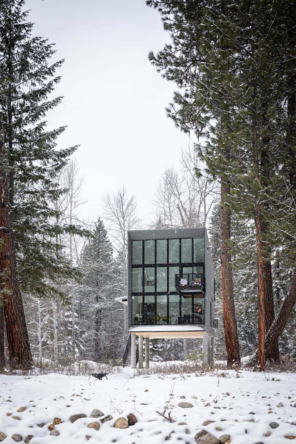Modern glass cabin on concrete piers in heavy snowfall surrounded by trees