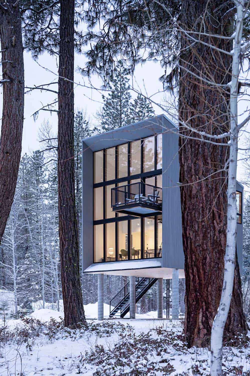Modern cabin with warm interior glow and mezzanine balcony seen through snowy birch trees