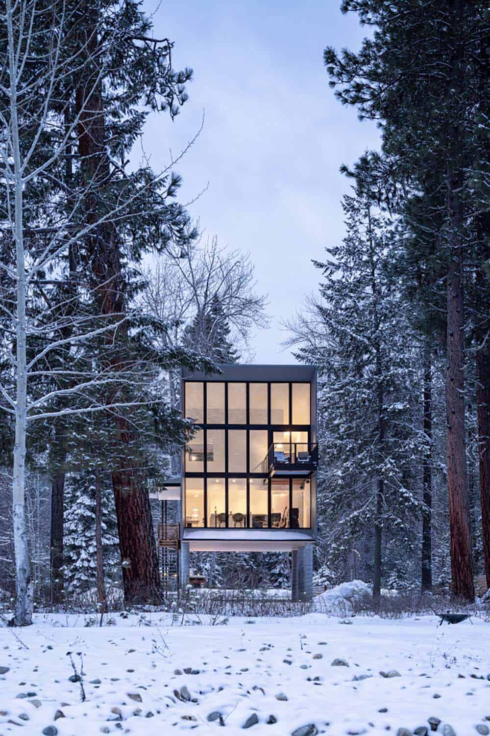Modern glass cabin glowing at dusk surrounded by snow-covered pine forest