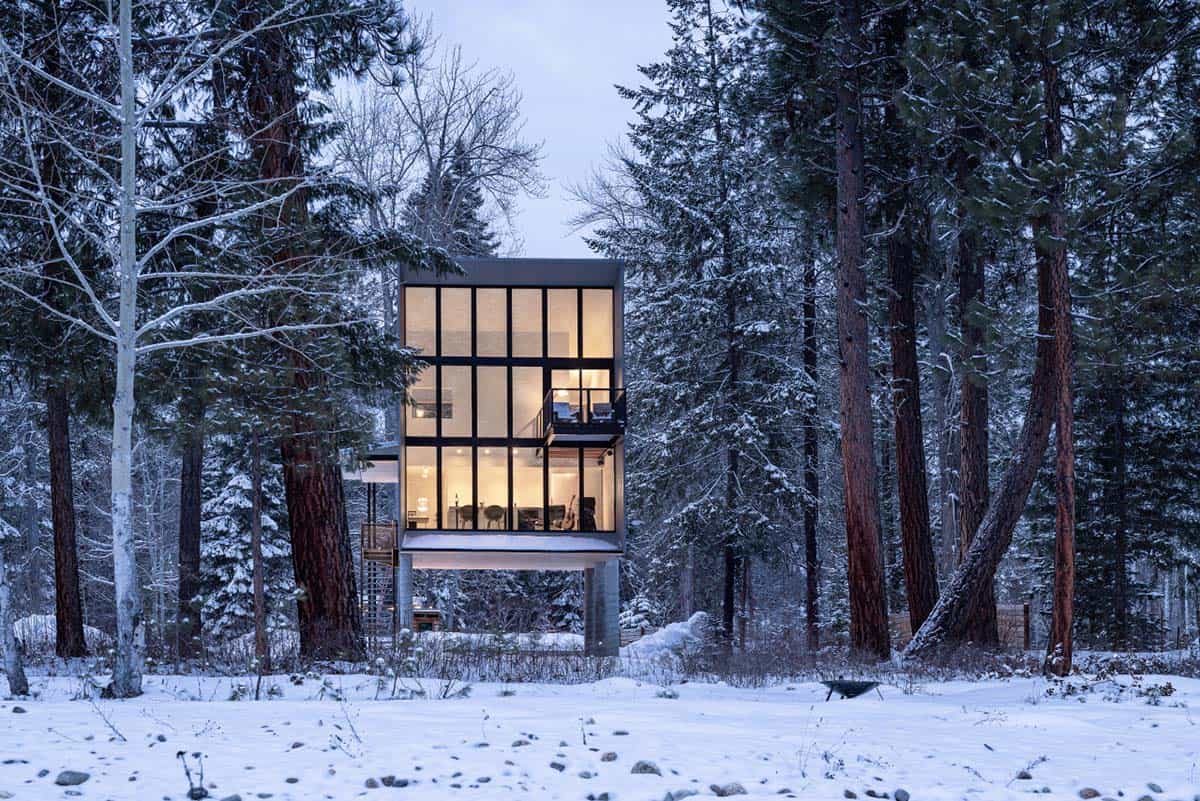 Modern cabin with full-height steel-framed windows glowing at blue hour in winter forest