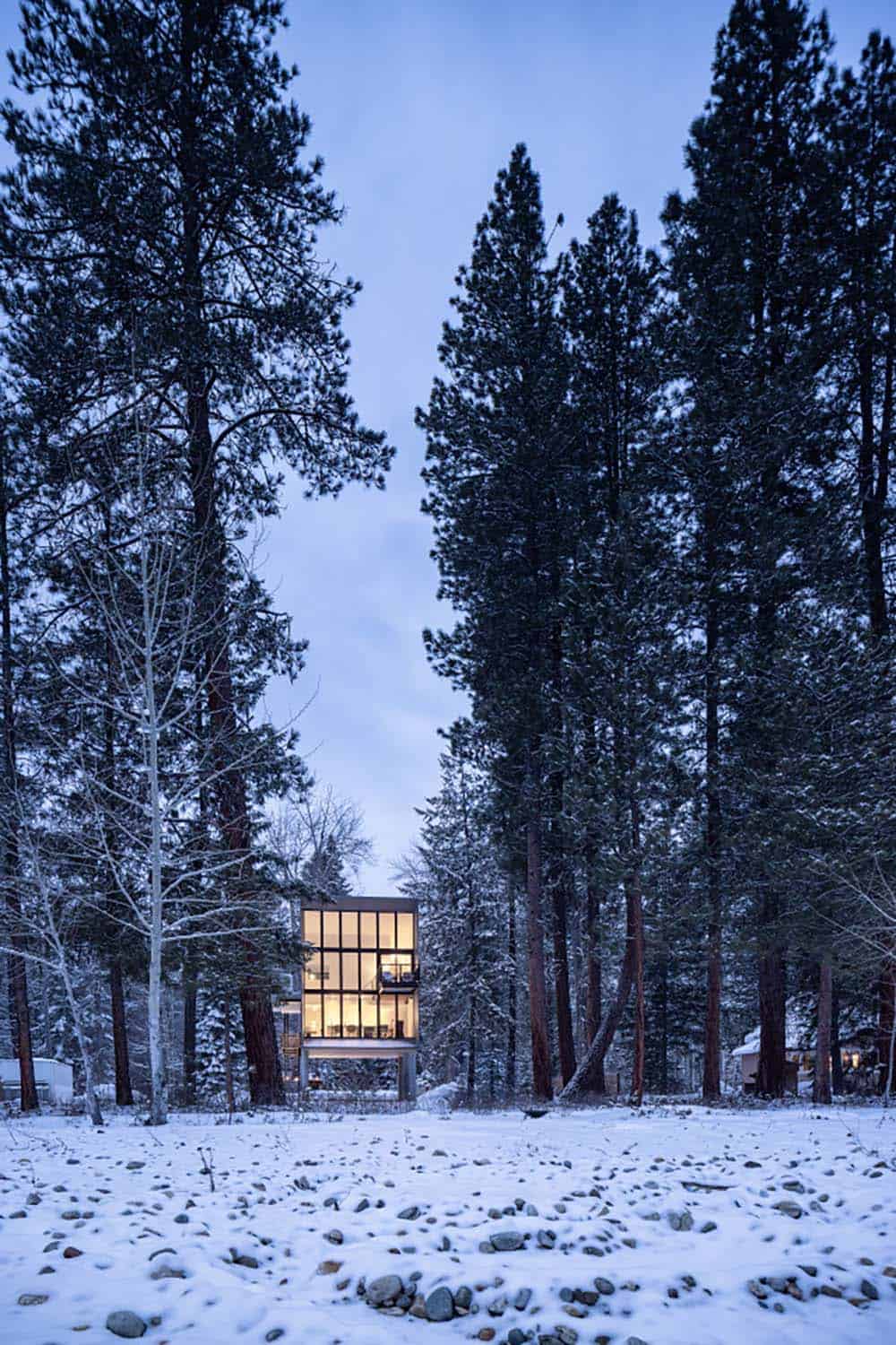 Distant winter dusk view of glowing modern cabin deep in snow-covered pine forest