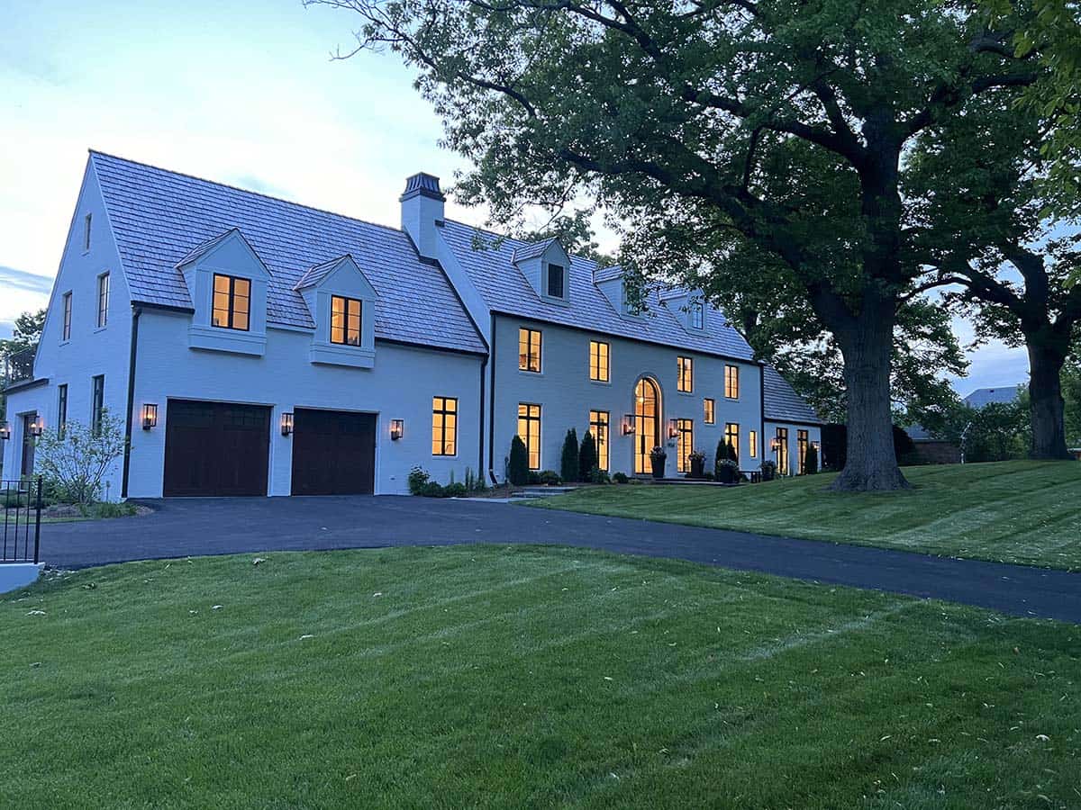 Twilight exterior view of a white brick Tudor-style manor with slate roof, dormers, and dual dark wood garage doors