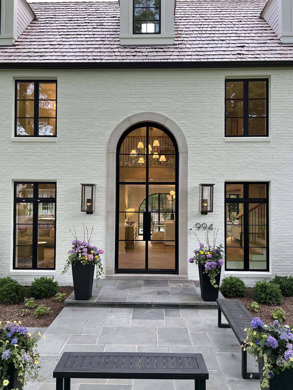 Close-up of arched glass front door flanked by black steel windows, stone surround, and flower-filled planters