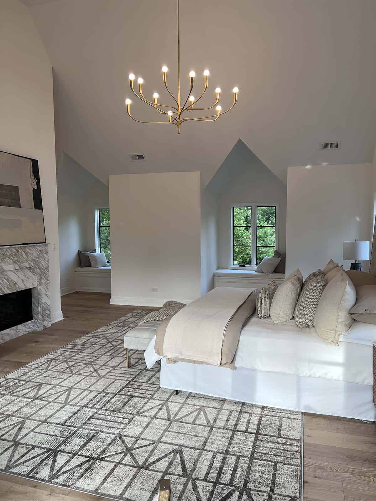 Vaulted primary bedroom with marble fireplace, gold sunburst chandelier, and twin dormer window seats