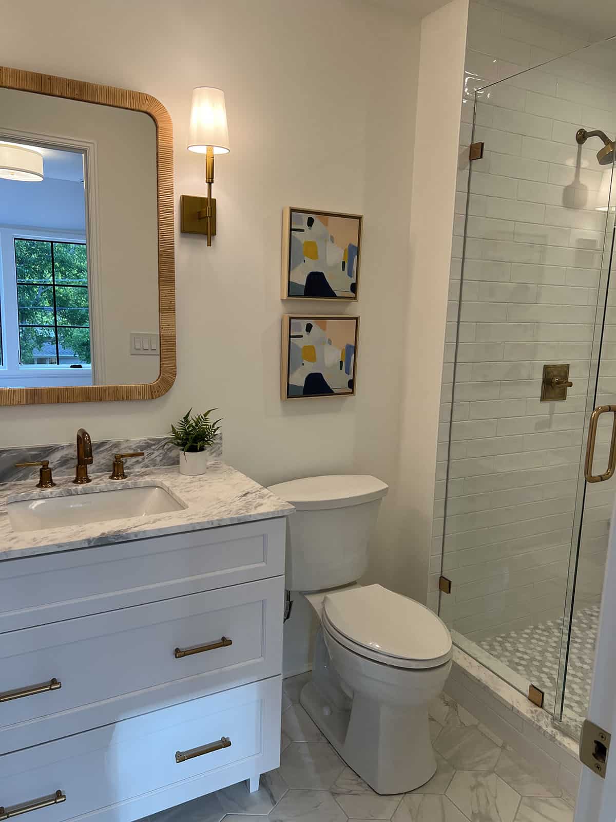 Small bathroom with rattan-framed mirror, marble vanity, brass hardware, and glass shower enclosure