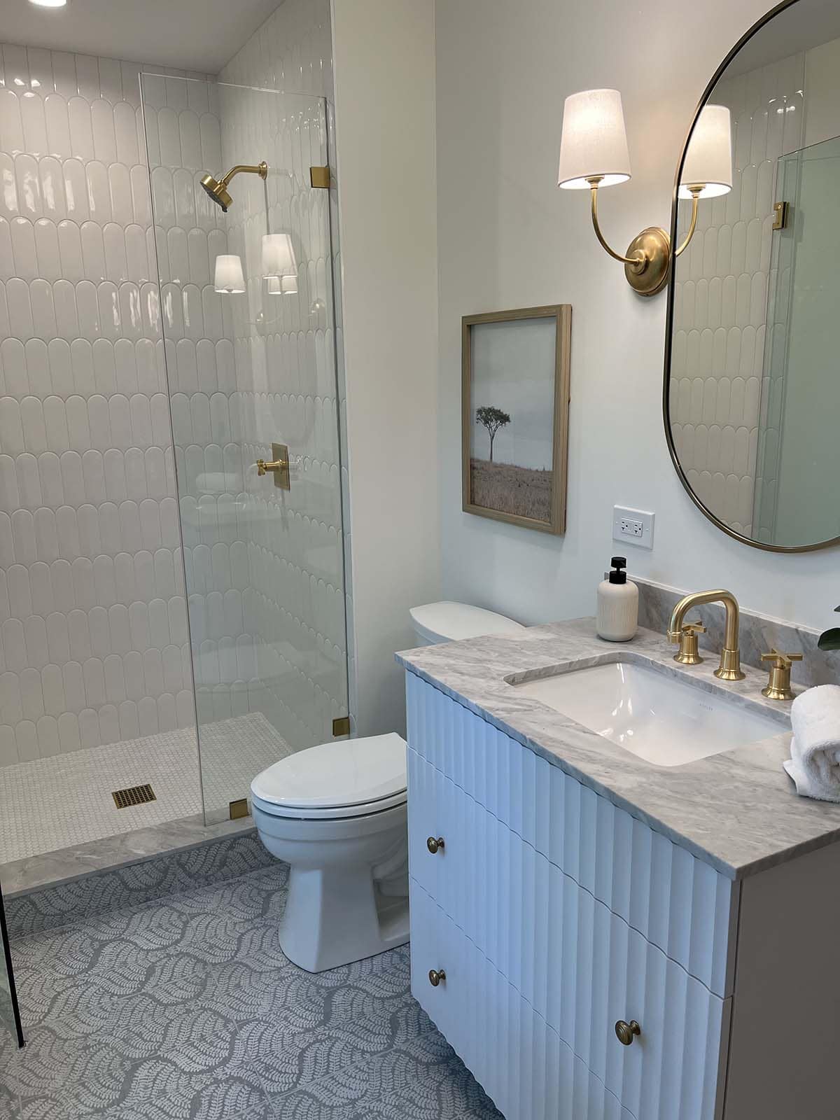 Bathroom with scalloped tile shower, round mirror, fluted blue vanity, and patterned tile floor