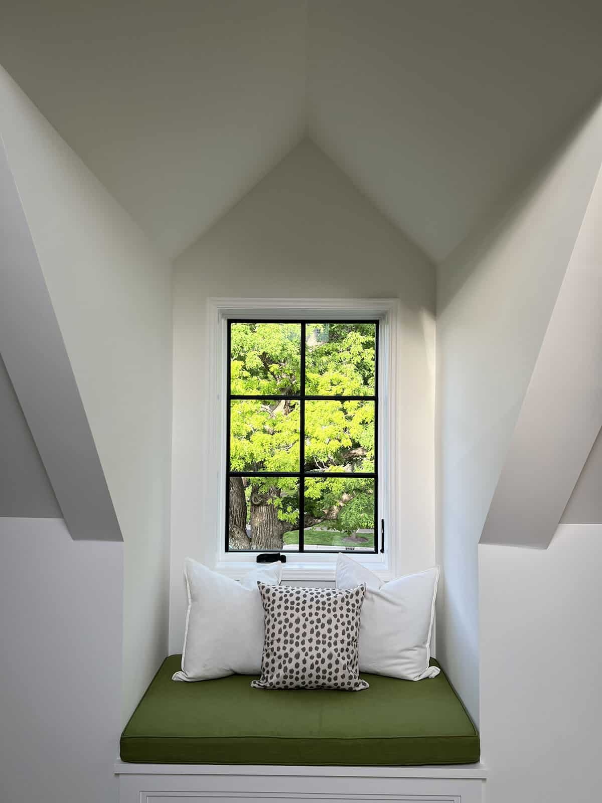 Dormer window nook with green cushioned bench seat and pillows overlooking leafy tree canopy