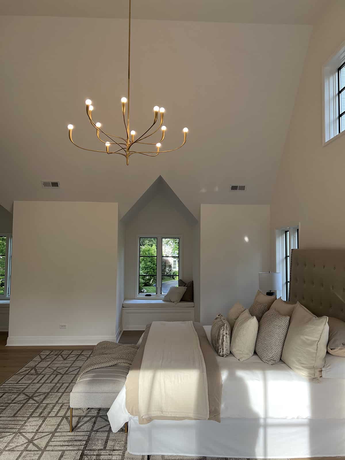 Wide view of vaulted primary bedroom showing tufted headboard, gold chandelier, and arched window nook