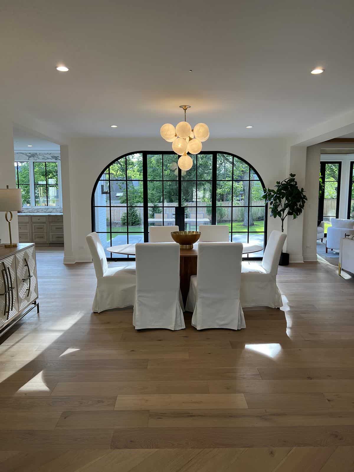 Open dining area with round wood table, white slipcovered chairs, and globe chandelier before arched window