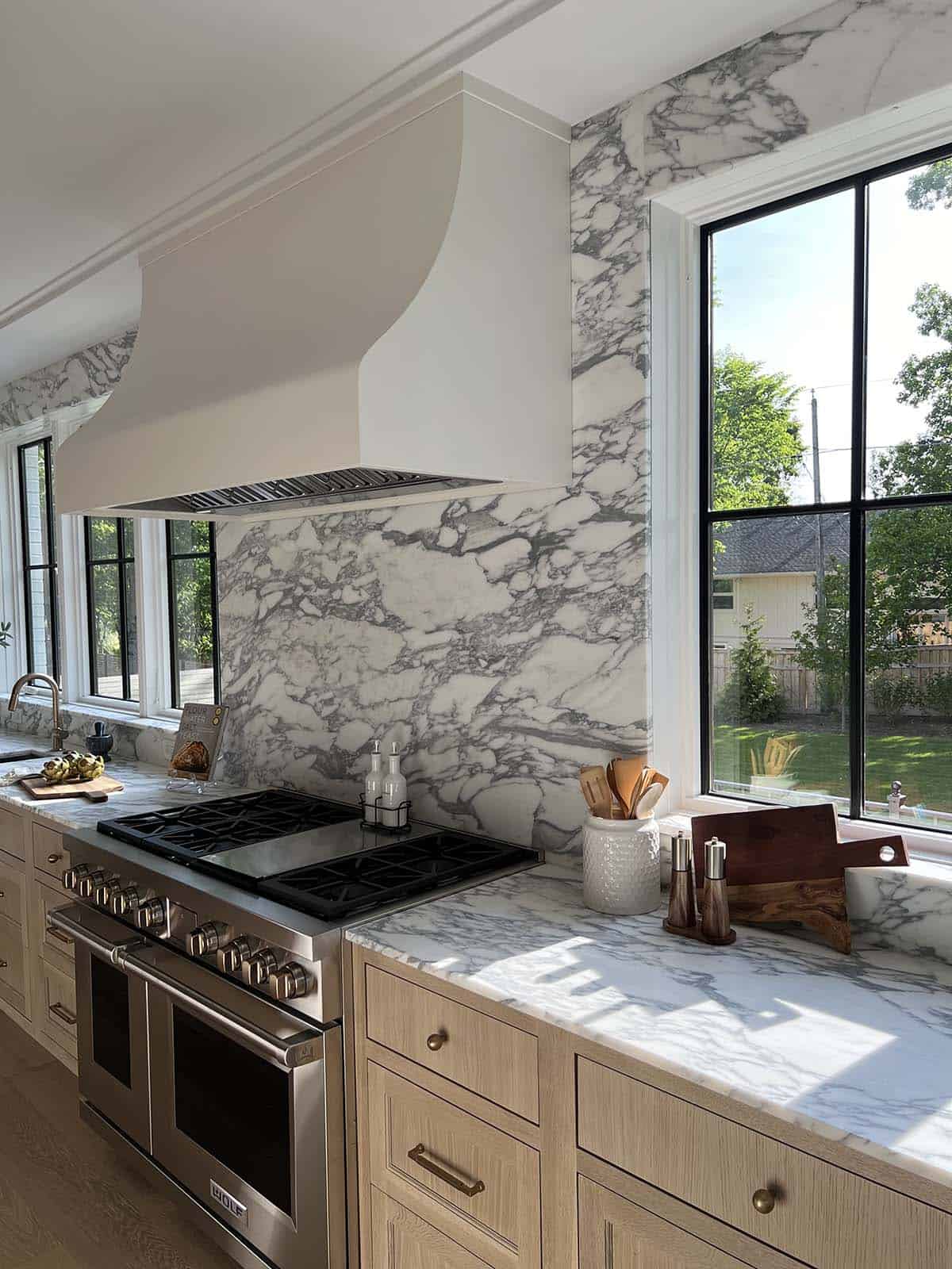 Angled view of range with marble backsplash, brass hardware, and steel window overlooking backyard