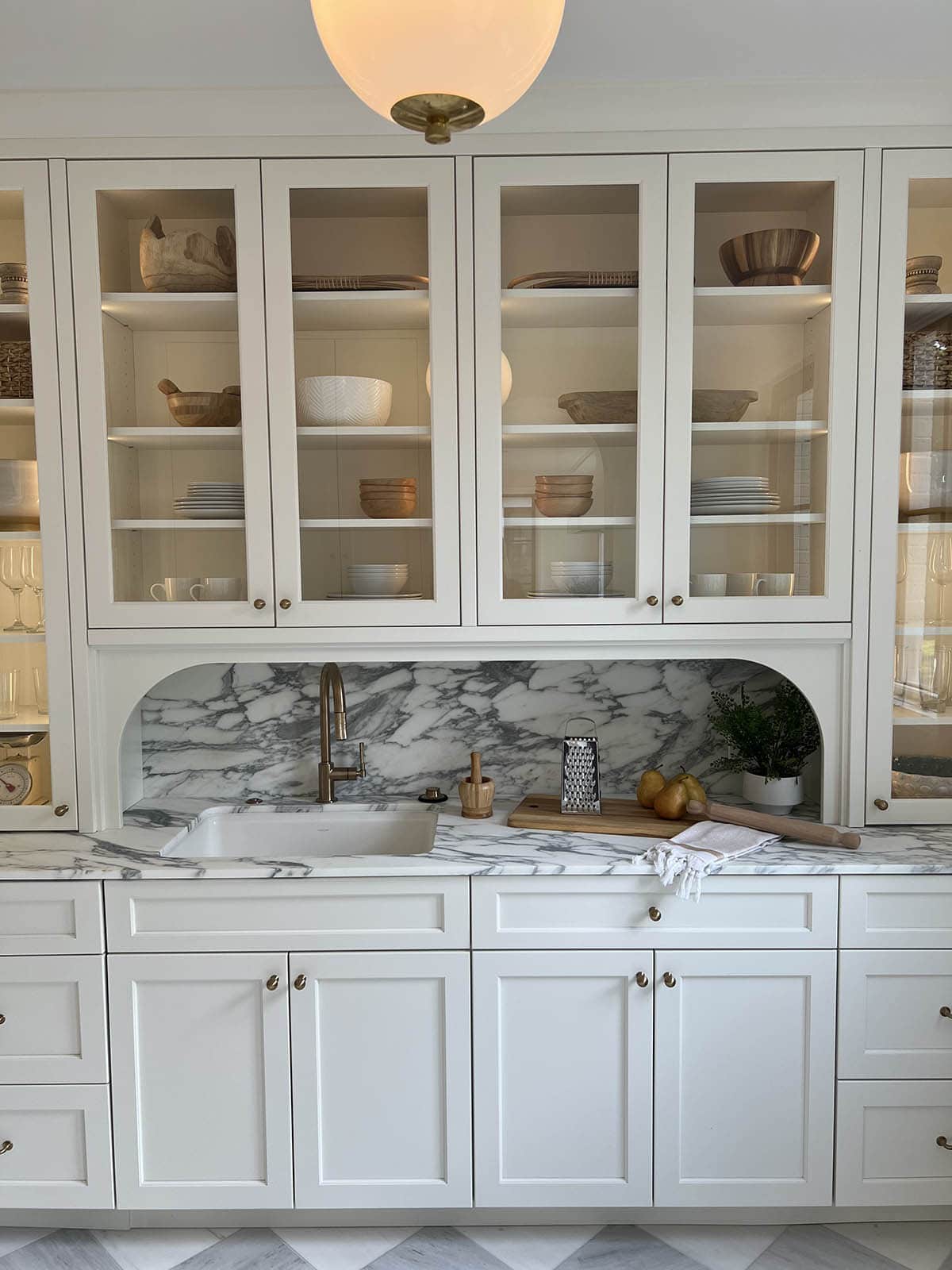 Butler's pantry with glass-front upper cabinets displaying dishware, marble backsplash niche, and brass faucet