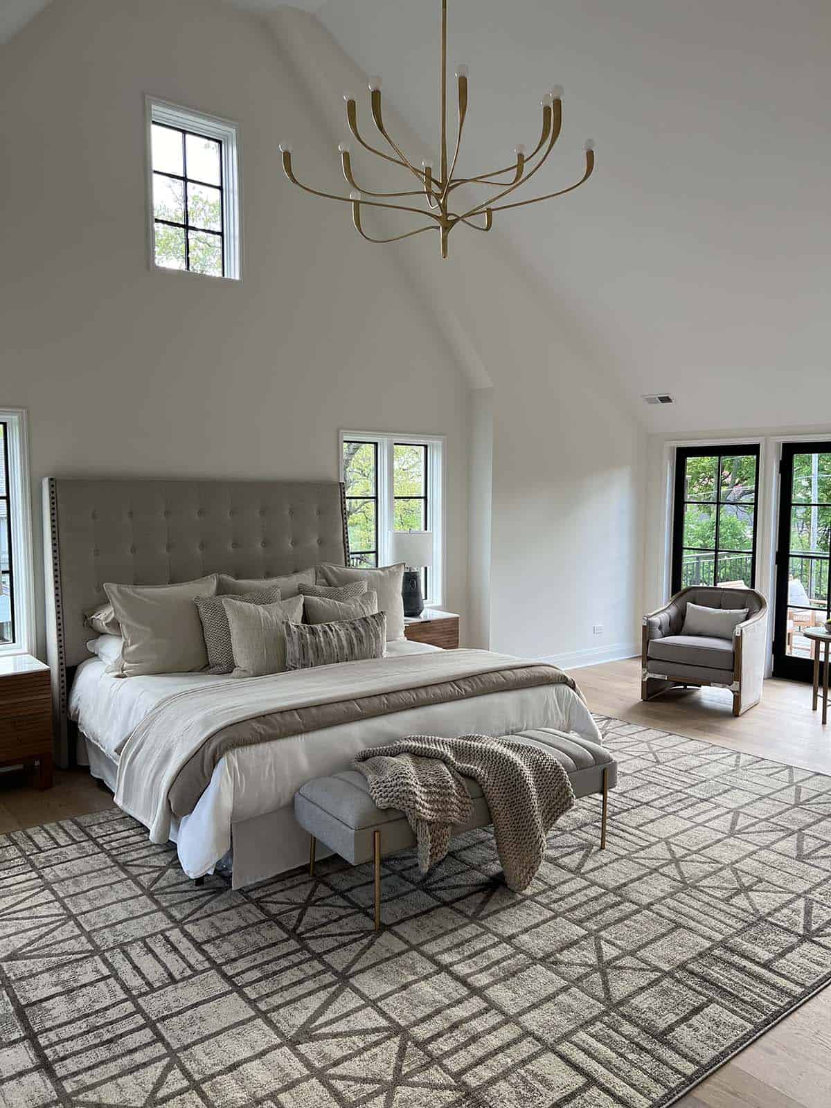 Primary bedroom with tufted gray headboard, geometric rug, and vaulted ceiling with brass chandelier