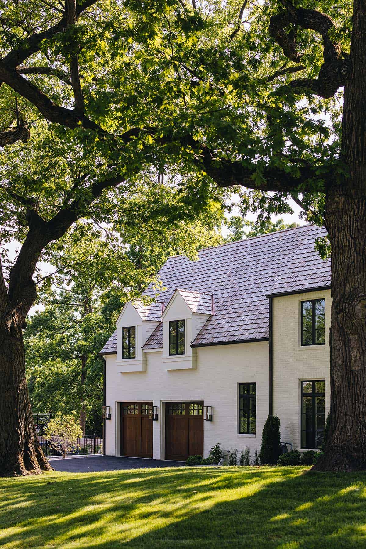 Side view of white brick garage with dormer windows, wood garage doors, framed by oak trees