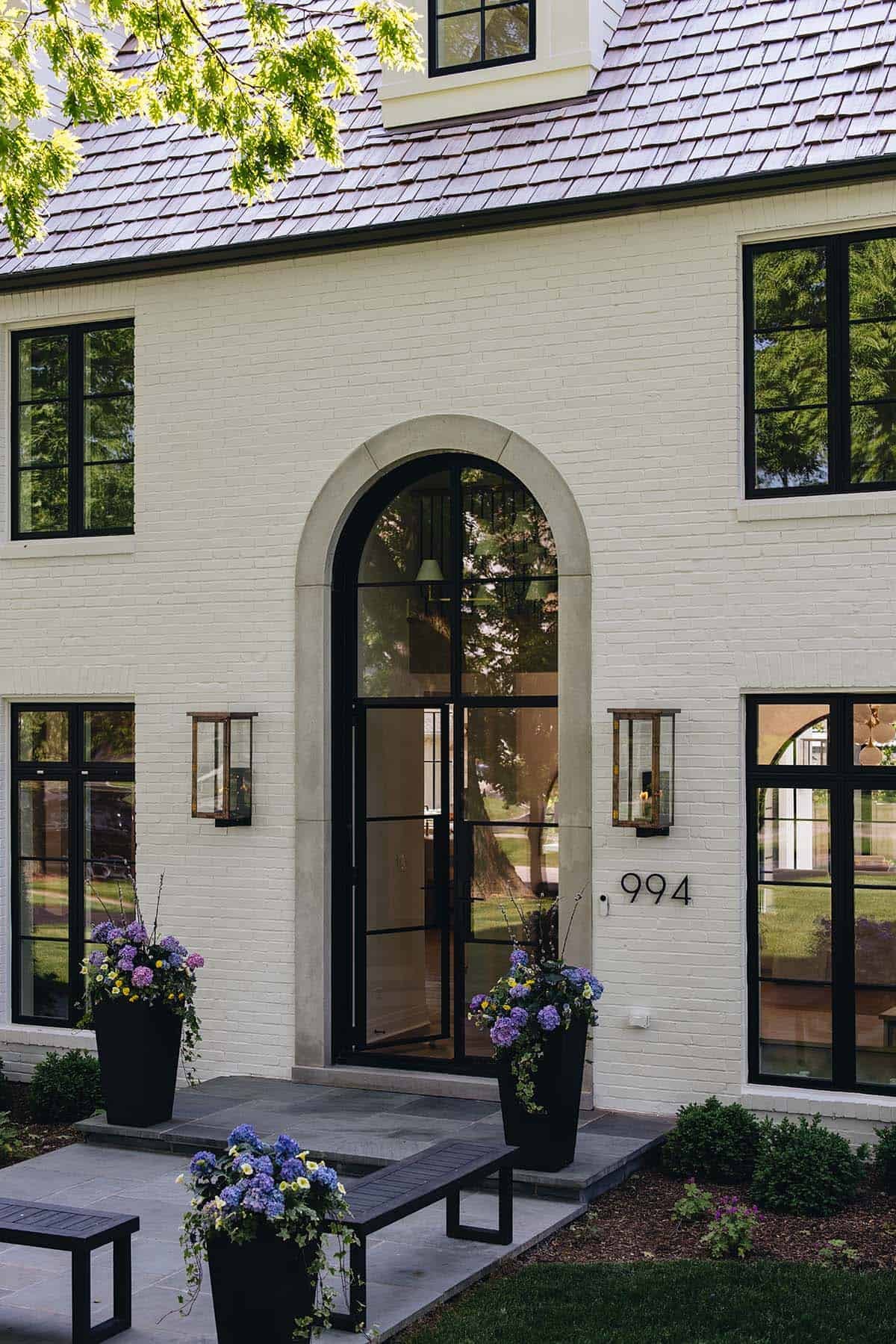 Close-up of arched steel front door with stone surround, sconces, and hydrangea planters at dusk