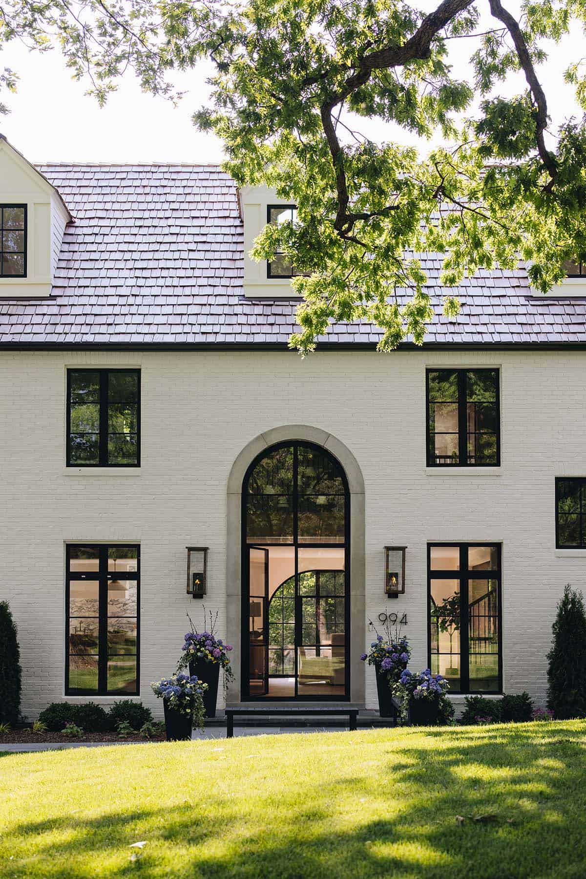 Sunlit view of arched front entry with open steel door, flower planters, and cedar shake roof