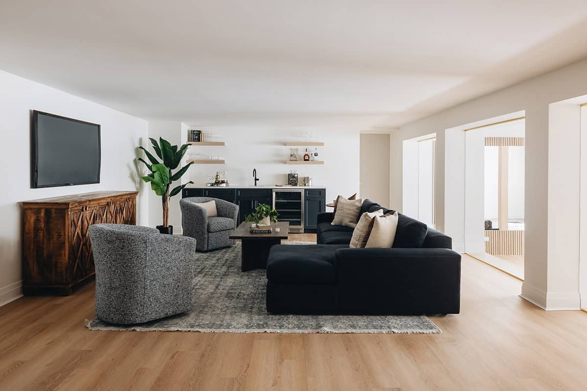 Basement lounge with navy sectional sofa, swivel chairs, wet bar with floating shelves, and wall-mounted TV