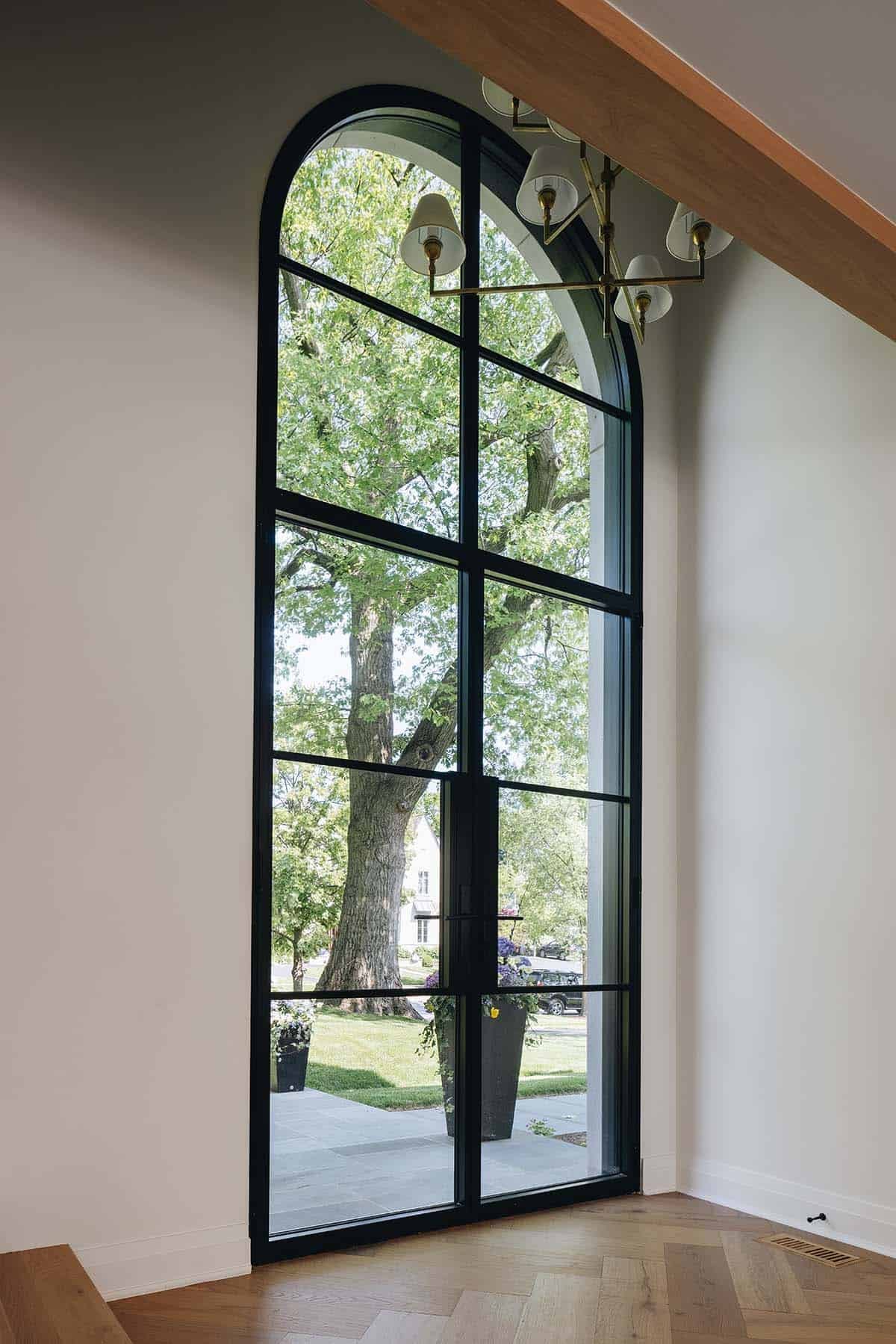 Tall arched steel-framed window and door with brass chandelier and tree view from foyer