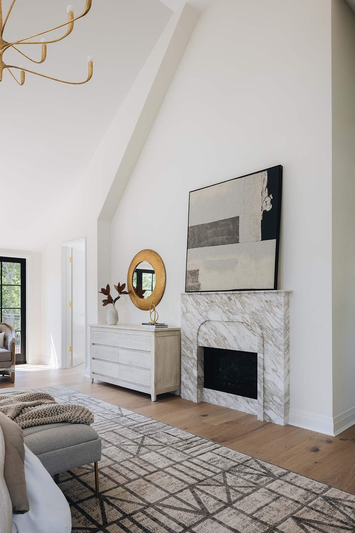 Vaulted bedroom corner with marble fireplace, abstract art, gold round mirror, and dresser
