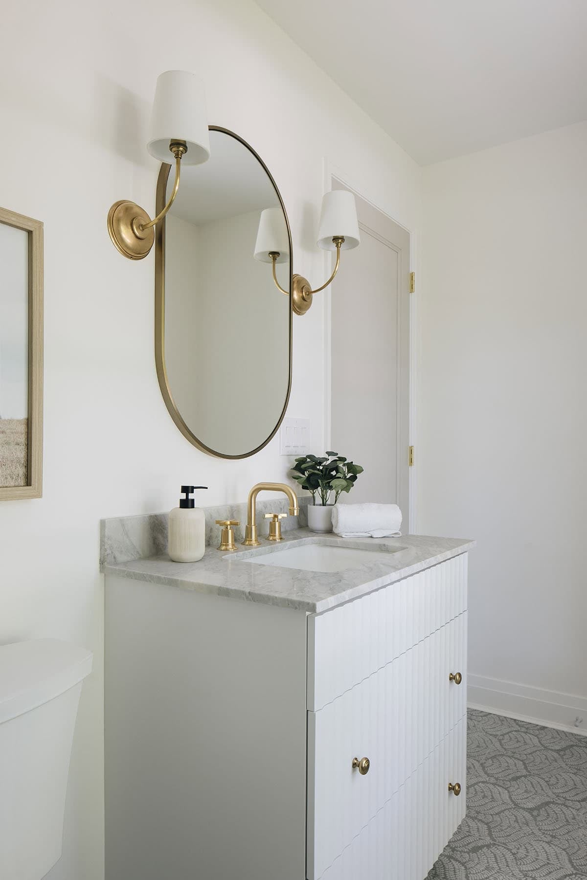 Bathroom vanity with oval brass mirror, twin sconces, and marble countertop with brass faucet