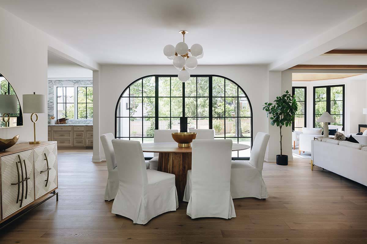 Open dining area with round wood table, white slipcovered chairs, and arched steel window