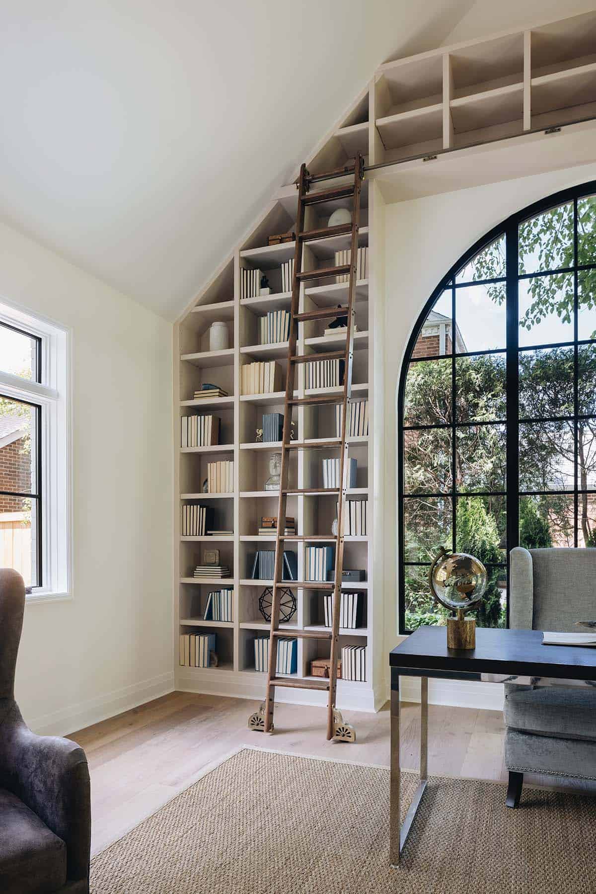 Home office library wall with rolling ladder, built-in bookshelves, and arched steel window