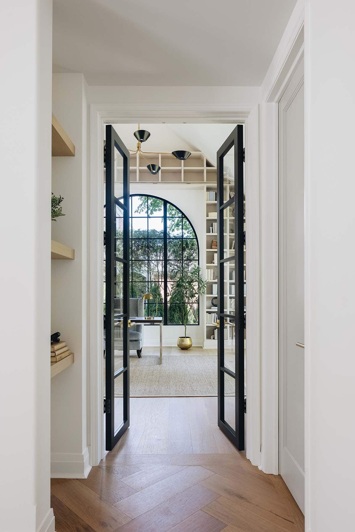 View through open glass French doors into home office with arched window and bookshelves