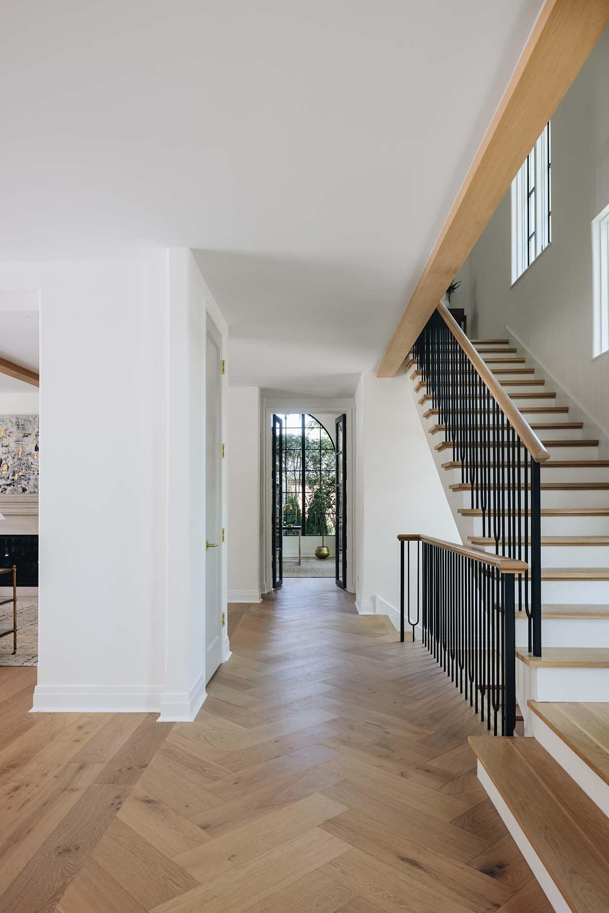 Herringbone-floored hallway with black staircase railing leading to arched glass door