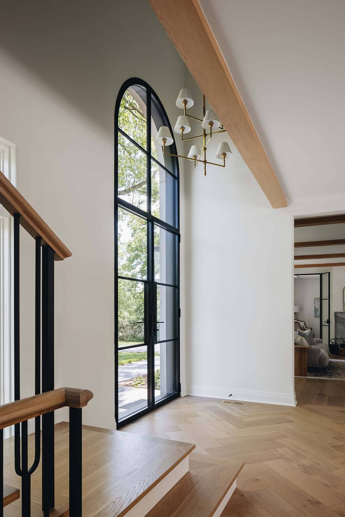 Tall arched black-framed door with brass chandelier and wood beam in foyer