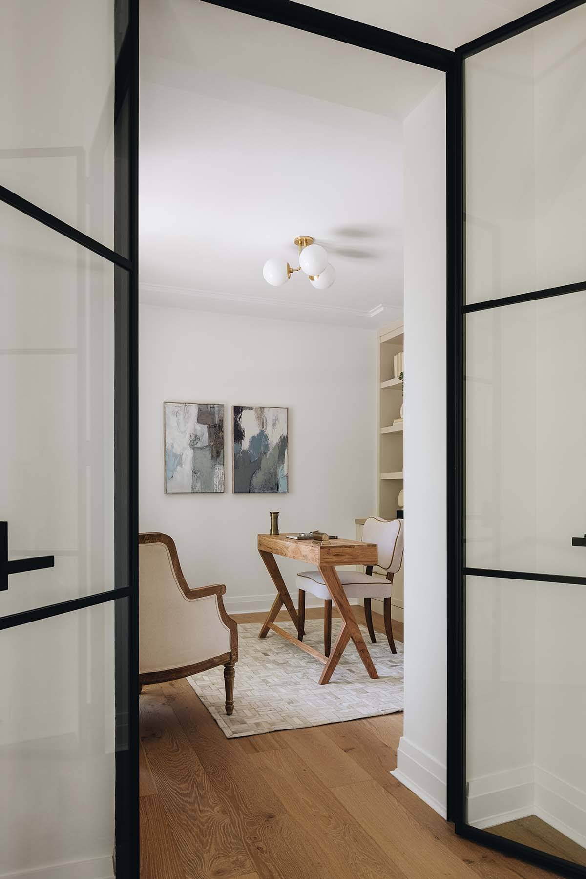 View through open black steel doors into home office with abstract art and wood desk