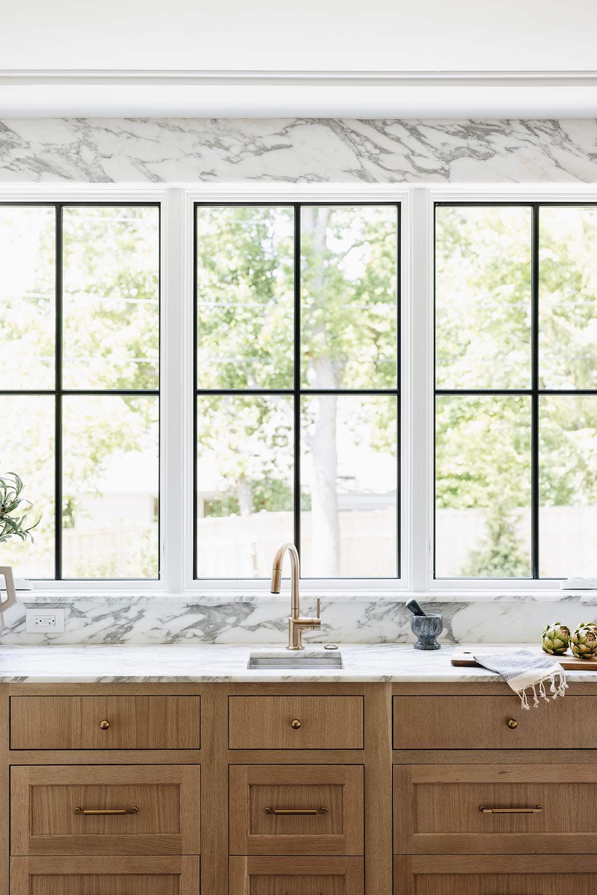 Close-up of three steel-framed windows above marble backsplash and oak drawer cabinetry