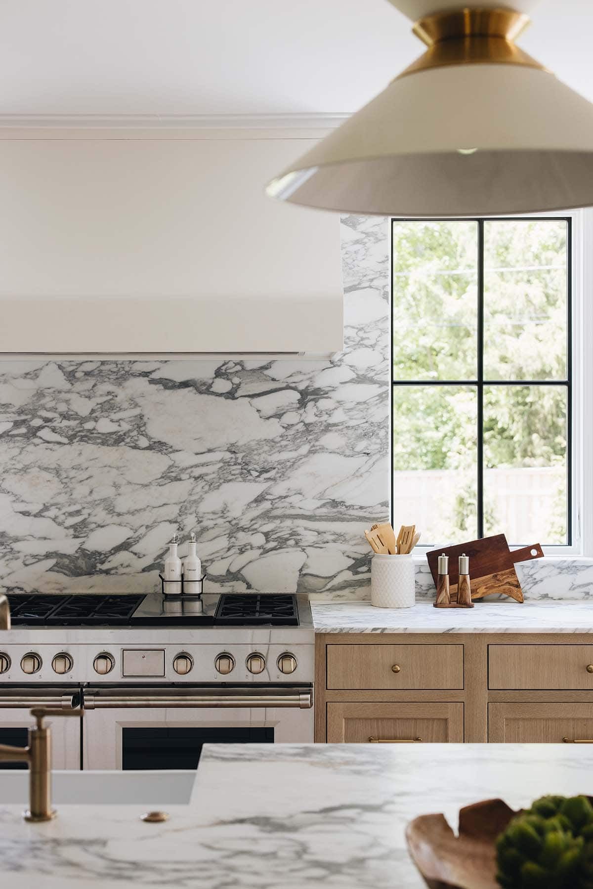Kitchen range detail with marble backsplash, brass pendant light, and wood cutting boards