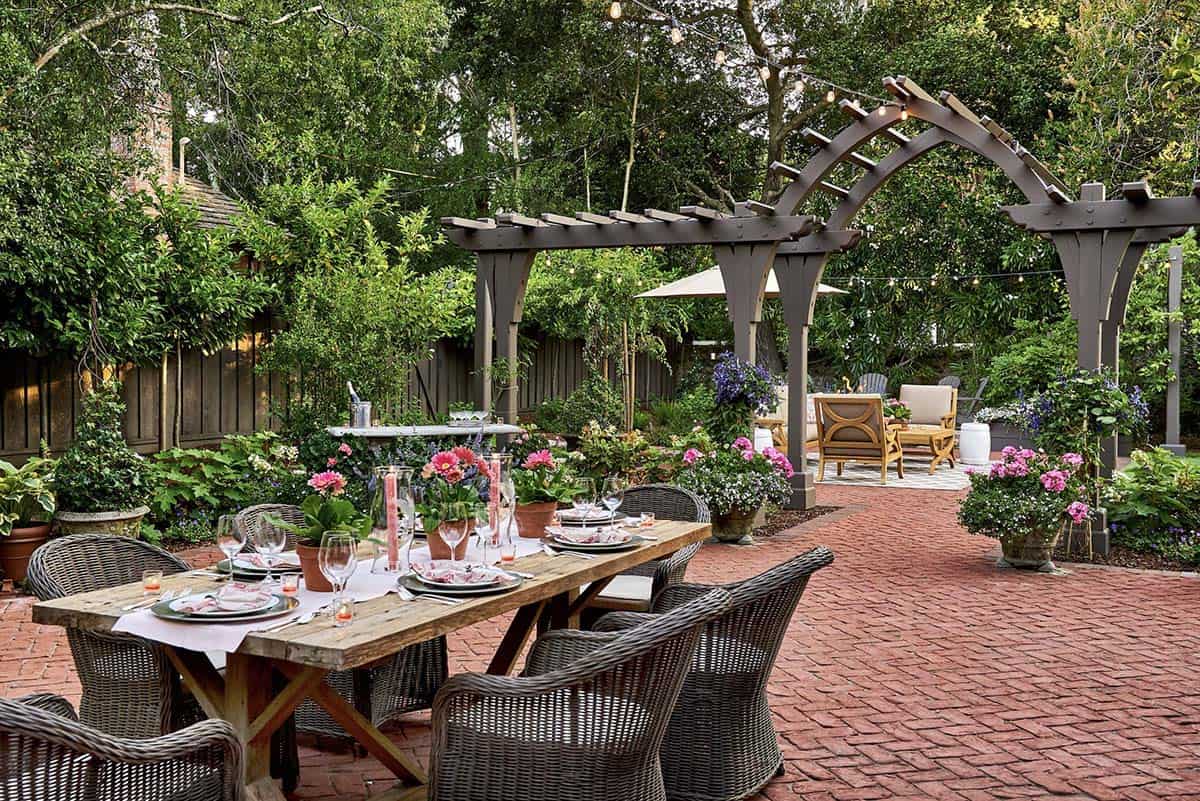 Formal outdoor dining table set with white linens, glassware, and pink flowers on a red brick patio, leading to an arched wooden pergola strung with string lights and a lounge seating area beyond
