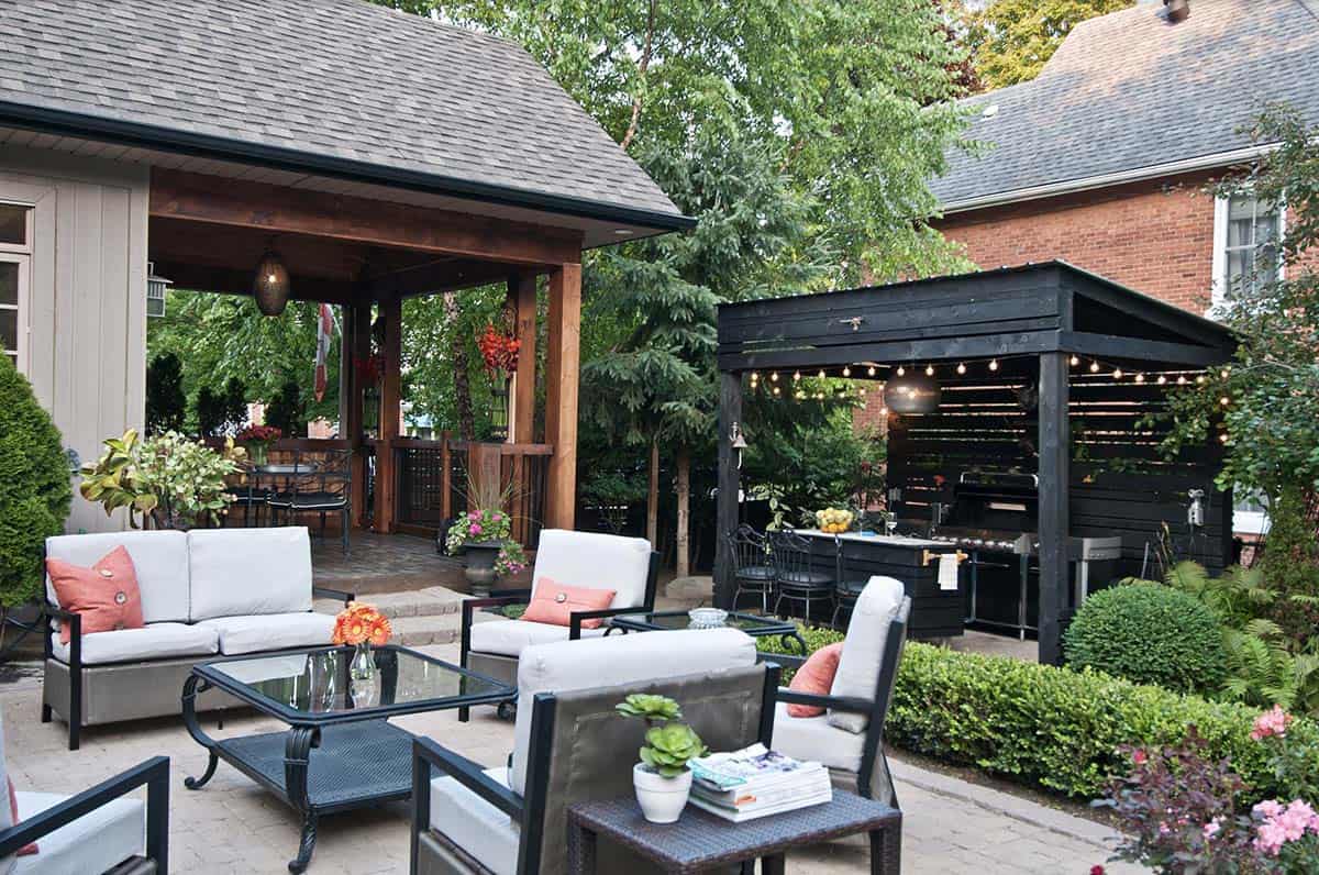 Backyard patio with gray cushioned lounge seating and glass coffee tables in the foreground, a covered wood pergola dining area to the left, and a black wood-clad outdoor kitchen structure with a built-in grill and string lights to the right