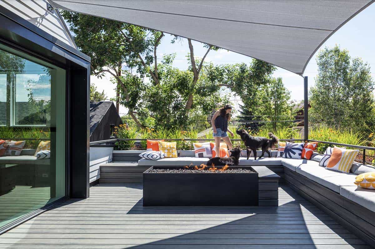 Modern rooftop deck with L-shaped built-in bench seating covered in colorful patterned pillows, a black rectangular fire pit, and a large gray shade sail overhead