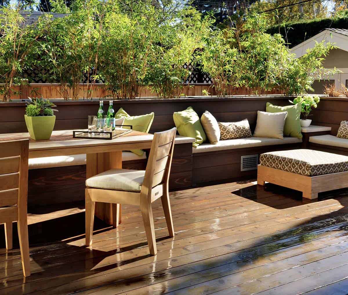 Wood deck with a built-in dark wood bench topped with cream and green patterned pillows, a teak dining table and chairs, and tall bamboo planted along the perimeter for privacy