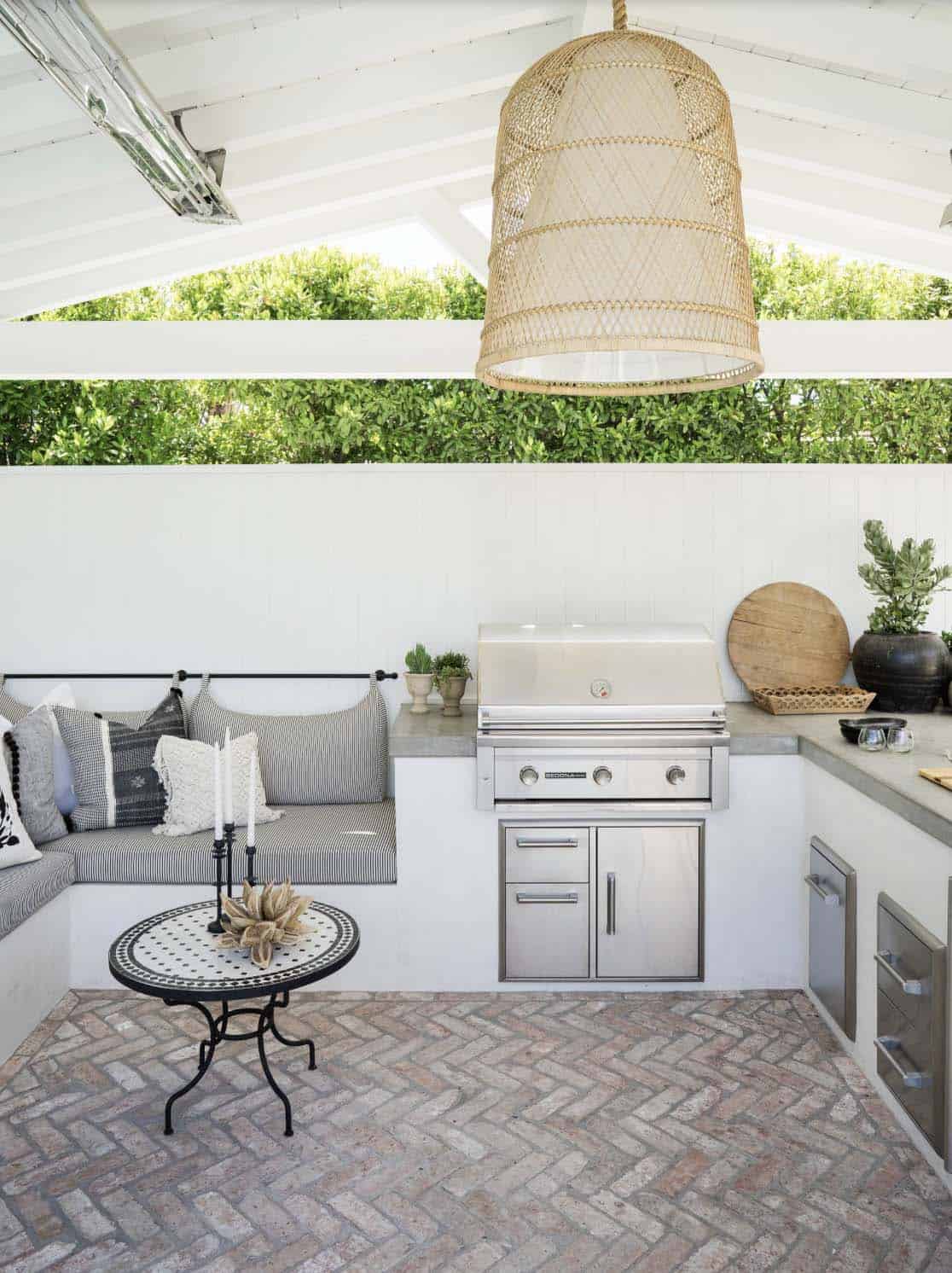 Covered outdoor kitchen nook with a built-in stainless steel grill, striped banquette seating with pillows, a mosaic side table, and a woven pendant light