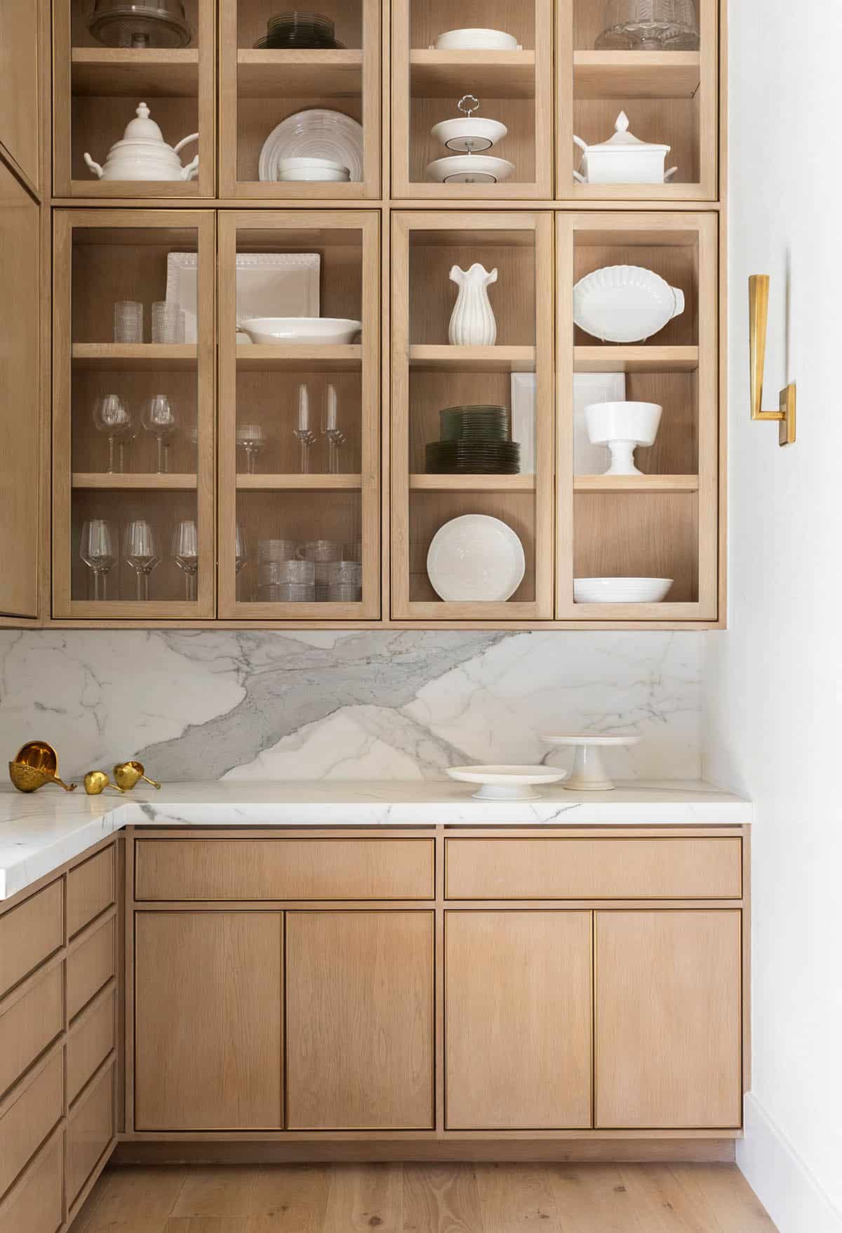 Glass-front oak cabinets displaying white dishware against marble backsplash