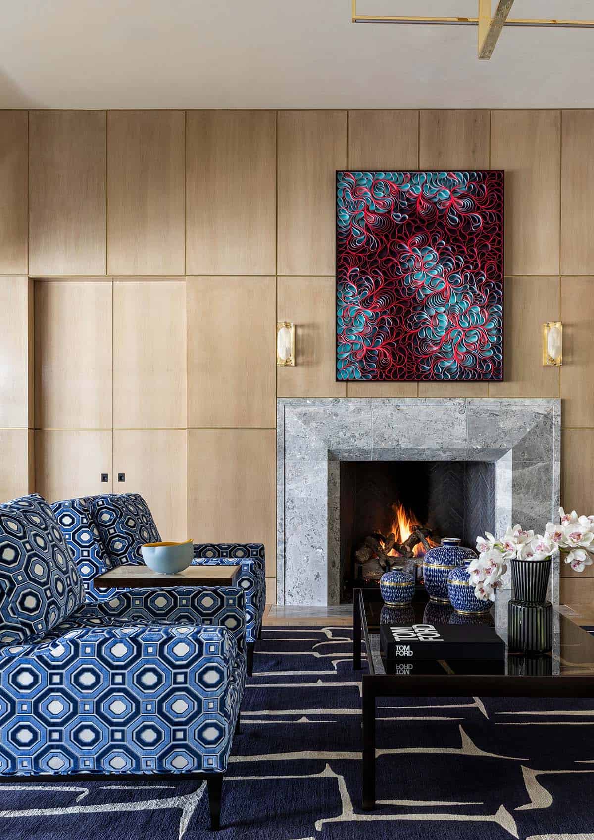 Wood-paneled living room with blue patterned chairs and colorful artwork above fireplace