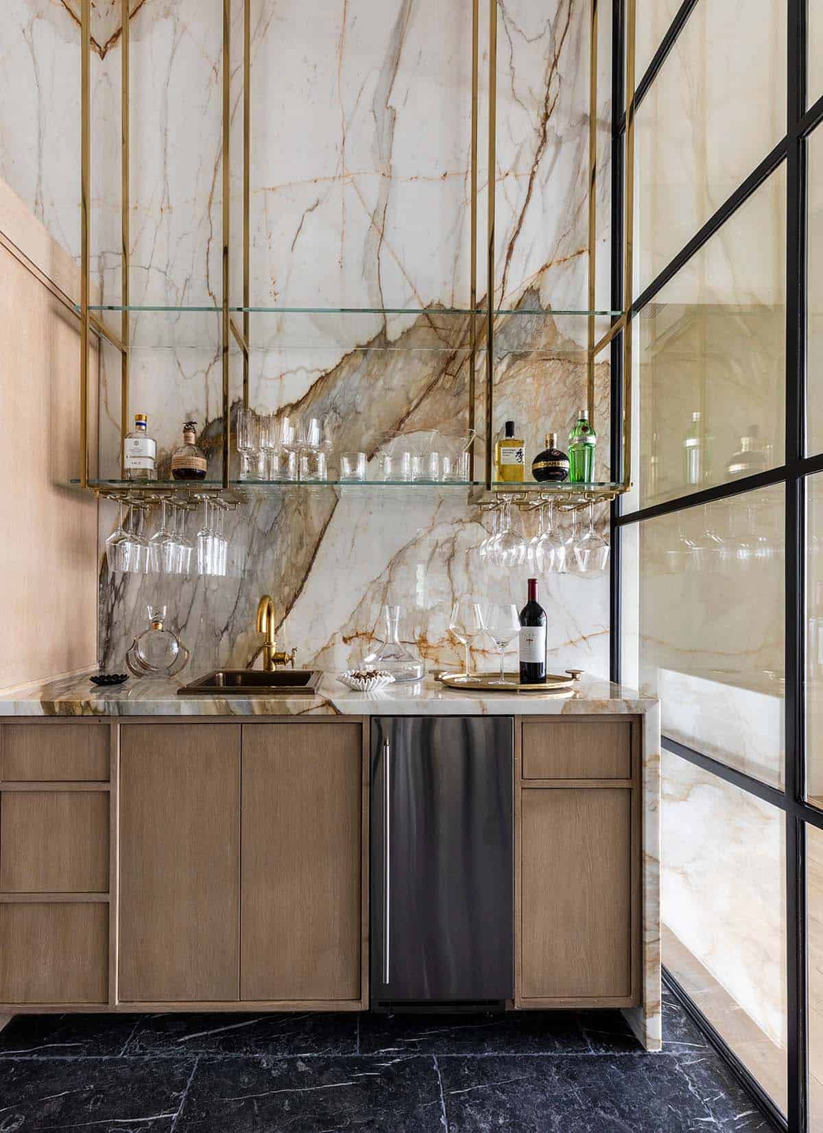 Wet bar with marble backsplash, brass fixtures, and glass shelving
