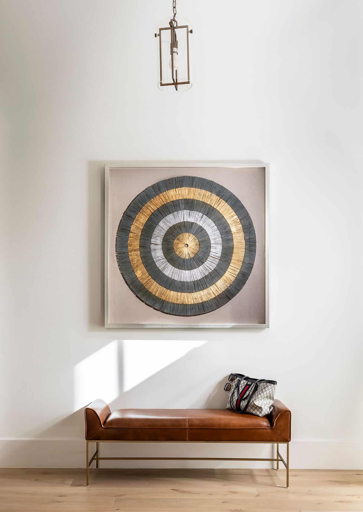 Circular gold and gray wall art above leather bench in entryway
