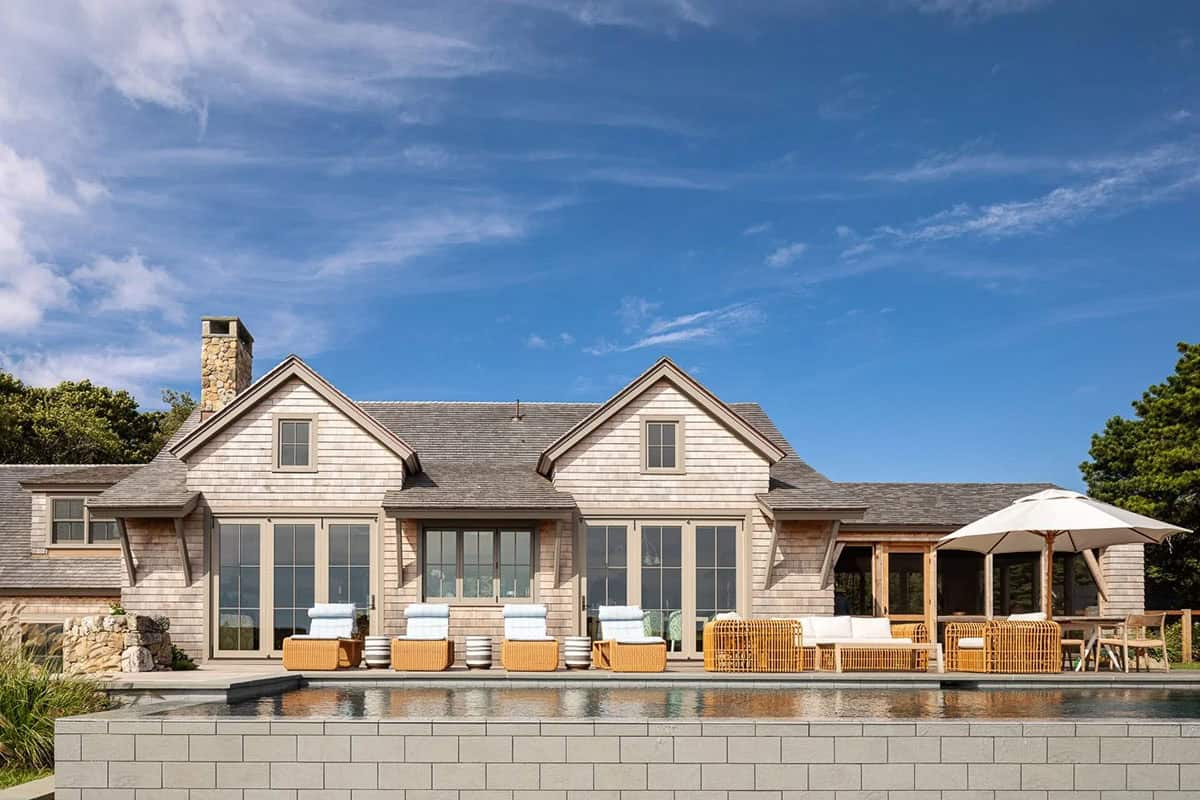 Shingle-style coastal home exterior with stone chimney, twin gables, and pool with wicker lounge chairs