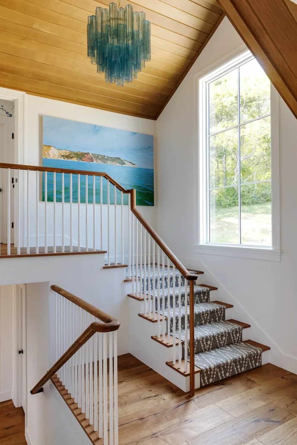 Bright stairwell with wood-paneled ceiling, blue glass chandelier, and framed coastal landscape painting